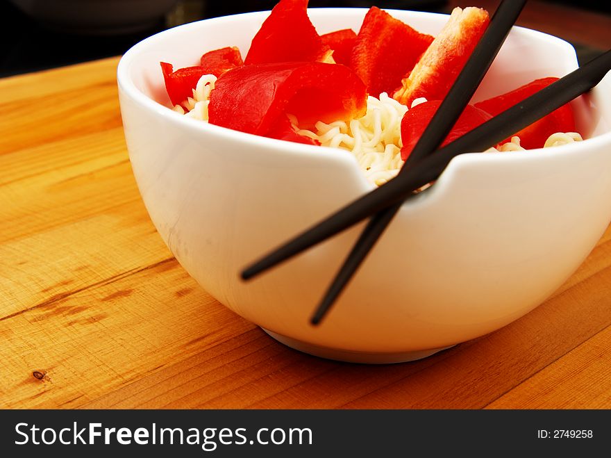 Chinece noodles with chop sticks in a bowl on wooden counter with some peppers in the noodles. Chinece noodles with chop sticks in a bowl on wooden counter with some peppers in the noodles