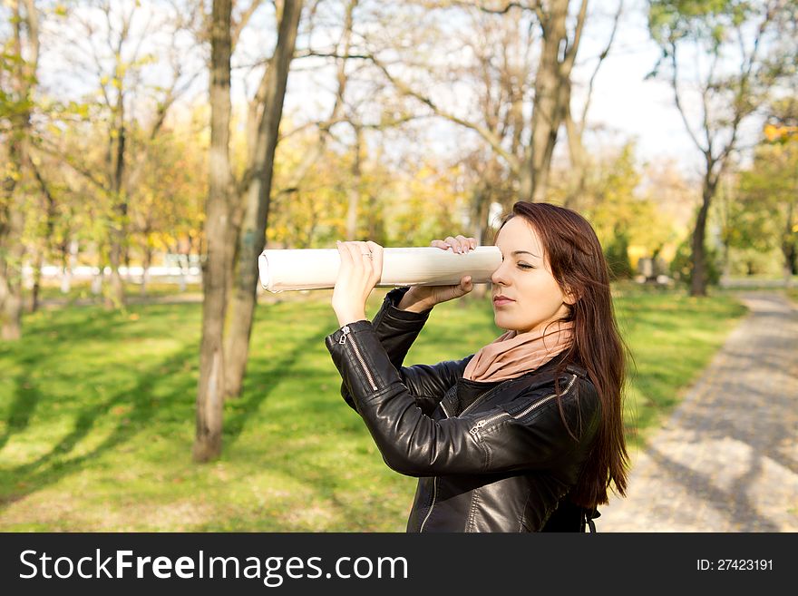 Woman using a rolled newspaper held up to her eye to spy over a distance standing outdoors in a wooded park. Woman using a rolled newspaper held up to her eye to spy over a distance standing outdoors in a wooded park