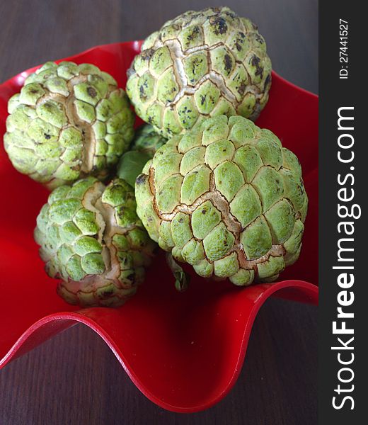 A bunch of sugar apple or Annona squamosa kept in a red bowl on a wooden table. A bunch of sugar apple or Annona squamosa kept in a red bowl on a wooden table