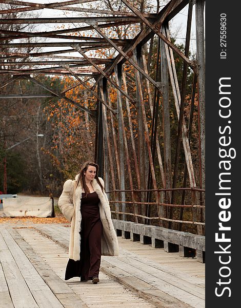 Pretty young woman wearing a long gown and fur coat goes for a walk over the old bridge in autumn. Pretty young woman wearing a long gown and fur coat goes for a walk over the old bridge in autumn.