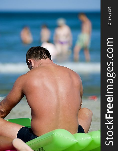 Man resting and sunbathing on the beach, people and sea in the background.