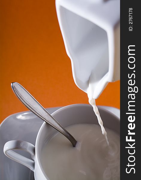 Close up of drooping milk in to a cup a dish and a spoon, in a orange background. Close up of drooping milk in to a cup a dish and a spoon, in a orange background