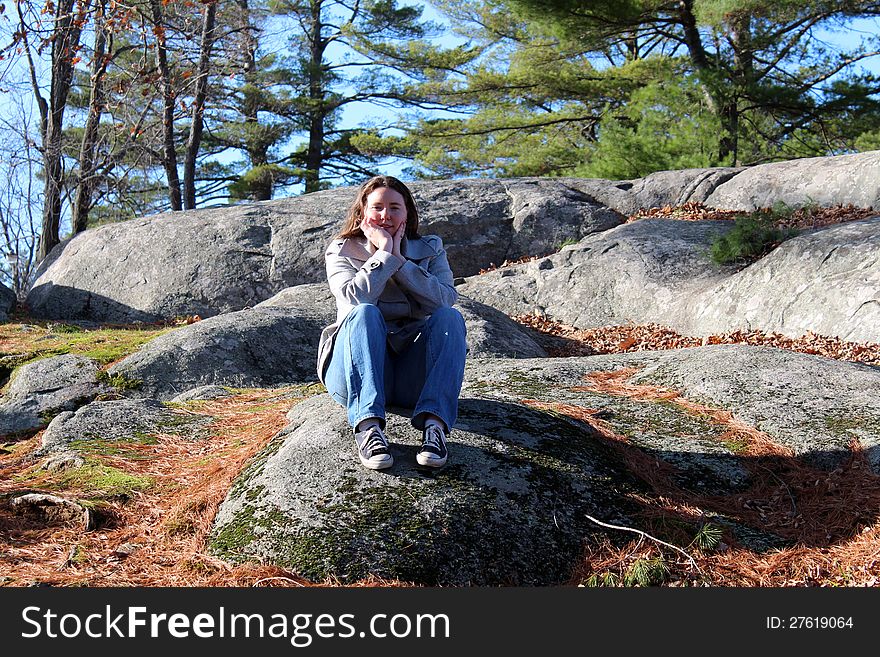 Young brunette woman dressed warmly sitting on rocks in the pine needle strewn woods. Young brunette woman dressed warmly sitting on rocks in the pine needle strewn woods.