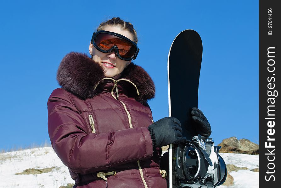 The young woman with a snowboard, in a helmet, in a ski glasses on the blue sky background