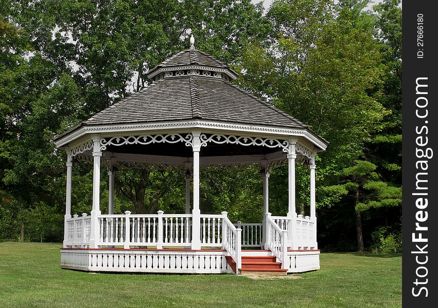 White wooden gazebo bandstand with a pointed shingled roof, in a park with grass and tall trees in the background. White wooden gazebo bandstand with a pointed shingled roof, in a park with grass and tall trees in the background.