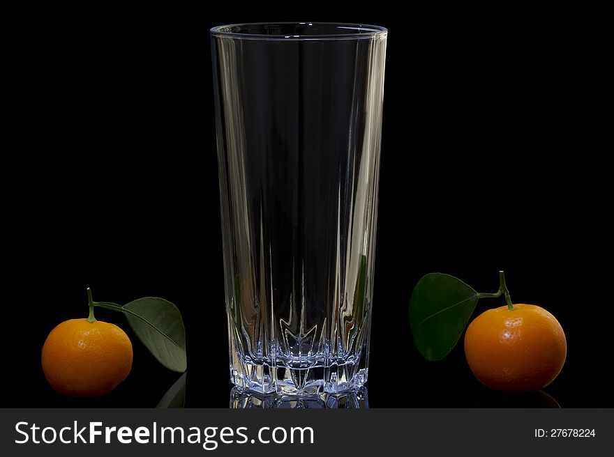 The composition of the three objects on a black background, one empty glass of clear glass and the right and left of the cup bright orange tangerines. Each mandarin on petiole have a green leaf. The composition of the three objects on a black background, one empty glass of clear glass and the right and left of the cup bright orange tangerines. Each mandarin on petiole have a green leaf.