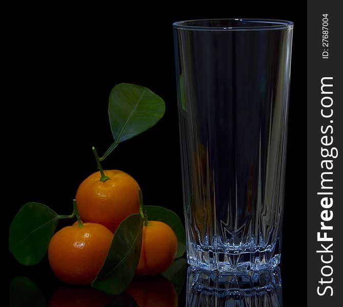 The composition of the objects on a black background, one high of cut glass and located beside him tangerines. Each mandarin on petiole have a green leaf. The composition of the objects on a black background, one high of cut glass and located beside him tangerines. Each mandarin on petiole have a green leaf.