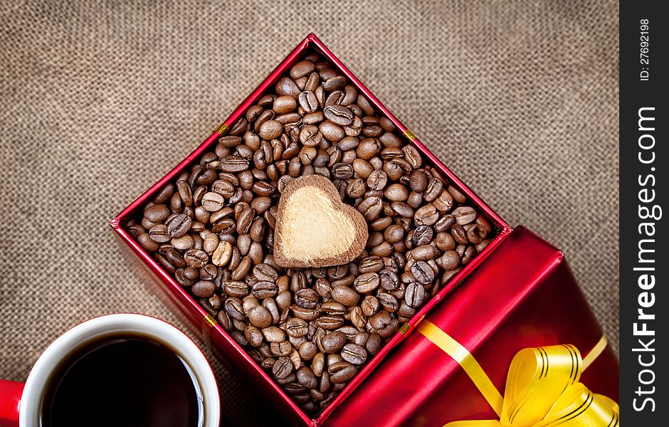 Coffee grains with heart shape cookie in Red box with yellow ribbon and mug with black coffee on textured brown background on Valentines day. Coffee grains with heart shape cookie in Red box with yellow ribbon and mug with black coffee on textured brown background on Valentines day