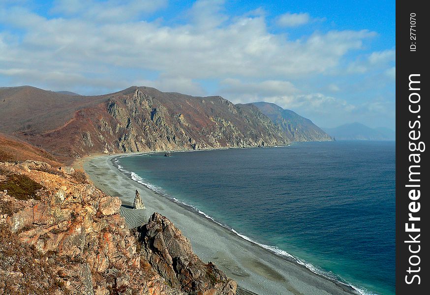 A cloudscape on sea: turquoise sea, grey beach, yellow-red capes and rocks and white clouds on azure sky. Autumn. Russian Far East, Primorye, Japanese sea, Tasovaya bay. A cloudscape on sea: turquoise sea, grey beach, yellow-red capes and rocks and white clouds on azure sky. Autumn. Russian Far East, Primorye, Japanese sea, Tasovaya bay.