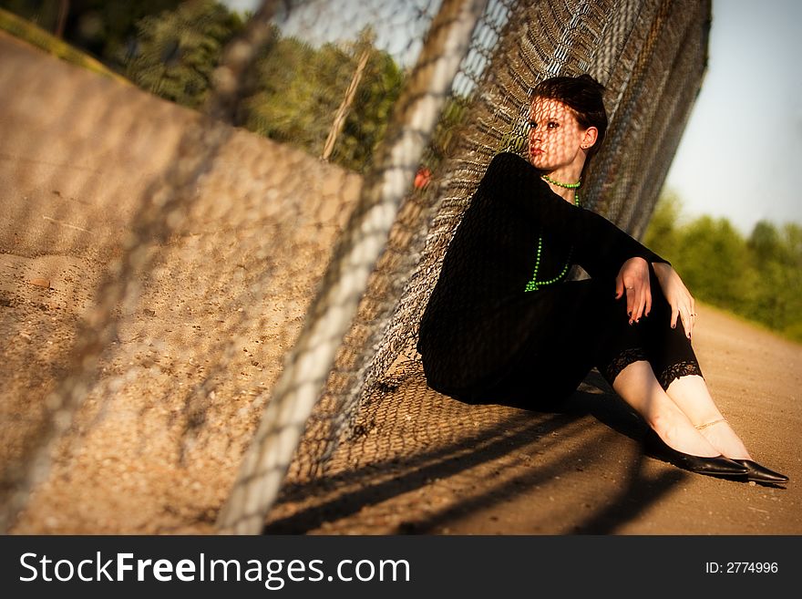 Attractive young lady sitting against a fence with a chain-link shadow criss-crossing her face. Attractive young lady sitting against a fence with a chain-link shadow criss-crossing her face.