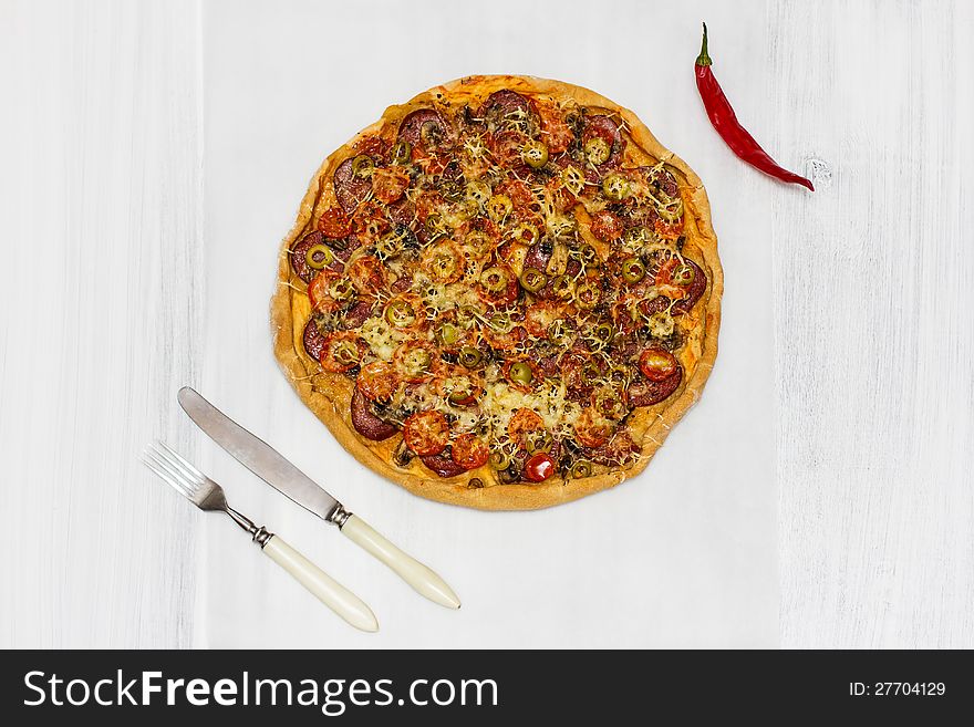 Pizza with salami and cherry tomatoes, with red chilli pepper and cutlery, on rusty white wood