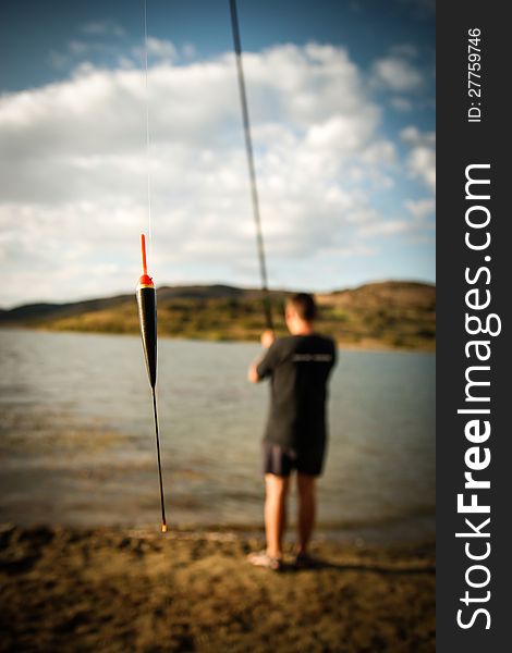 Lakeside fishing, focus on floater hanging from the line, the background is out of focus having the fisherman standing with the fishing rod facing away toward the lake