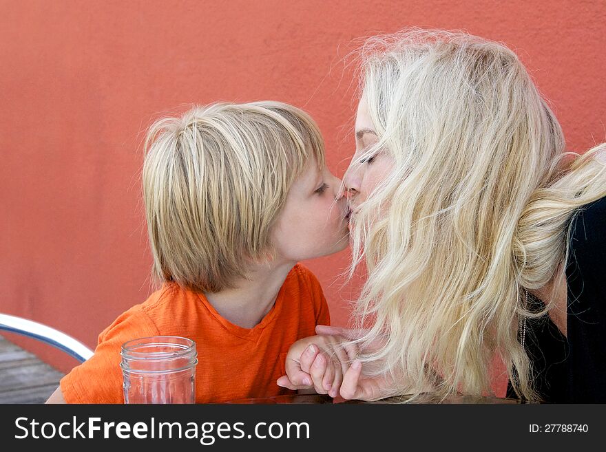 Beautiful blond mother kissing her preschool son against orange background. Beautiful blond mother kissing her preschool son against orange background