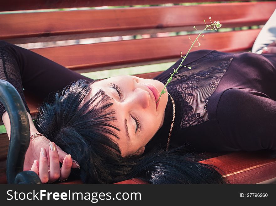 Young woman enjoying the sun, lying on a bench, with closed eyes and a flower in her lips. Young woman enjoying the sun, lying on a bench, with closed eyes and a flower in her lips