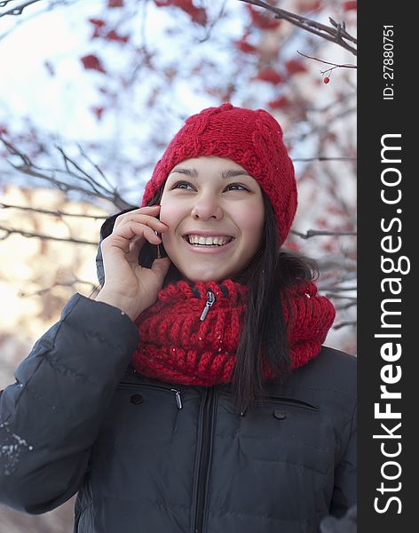 The young girl in a red knitted cap and a red scarf speaks by phone on the winter street. The young girl in a red knitted cap and a red scarf speaks by phone on the winter street