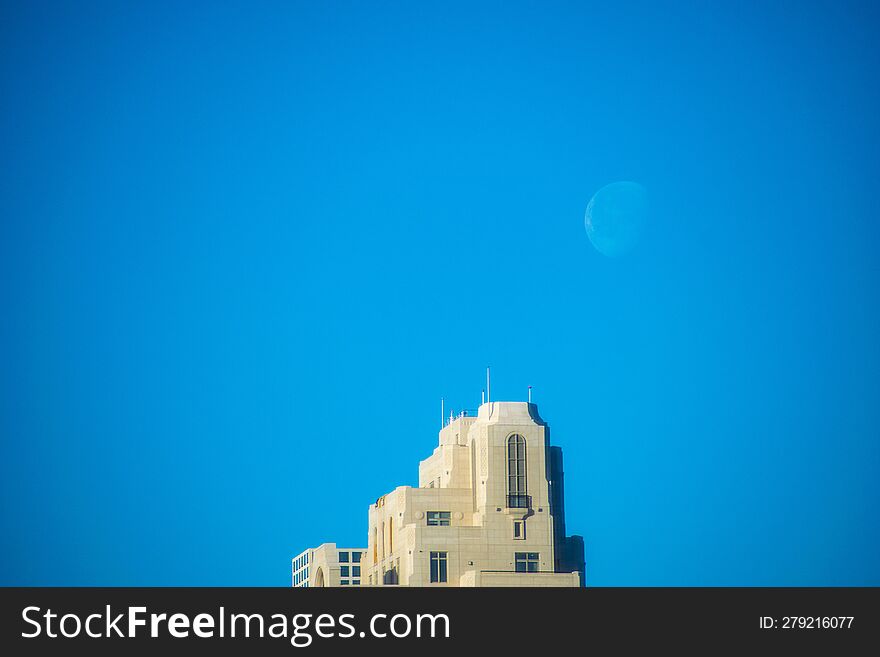 A bright white crescent moon rises over a blocky tan stone high rise skyscraper building with a true blue sky