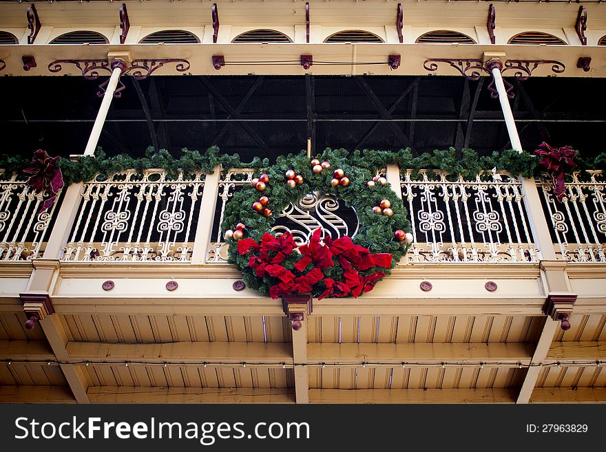 Christmas wreath hung on a late 1800s wrought iron & wood walkway. The wreath is made of green foliage with a red bow & gold decorations throughout. Evergreen garland lines the walkway handrail. Christmas wreath hung on a late 1800s wrought iron & wood walkway. The wreath is made of green foliage with a red bow & gold decorations throughout. Evergreen garland lines the walkway handrail.