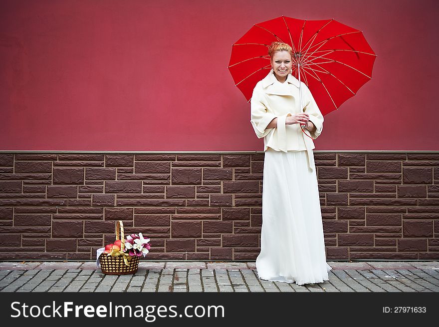 Woman in white coat with red umbrella and basket for picnic near burgundy walls. Woman in white coat with red umbrella and basket for picnic near burgundy walls