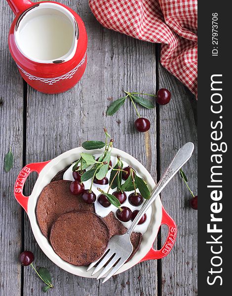 Chocolate Pancakes with Vanilla Cream and Cherries with a milk pot and checked napkin on a wooden surface