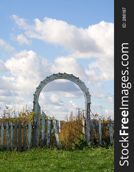 A dilapidated picket fence and gate lead to blue skies and grassy pastures beyond. A dilapidated picket fence and gate lead to blue skies and grassy pastures beyond.