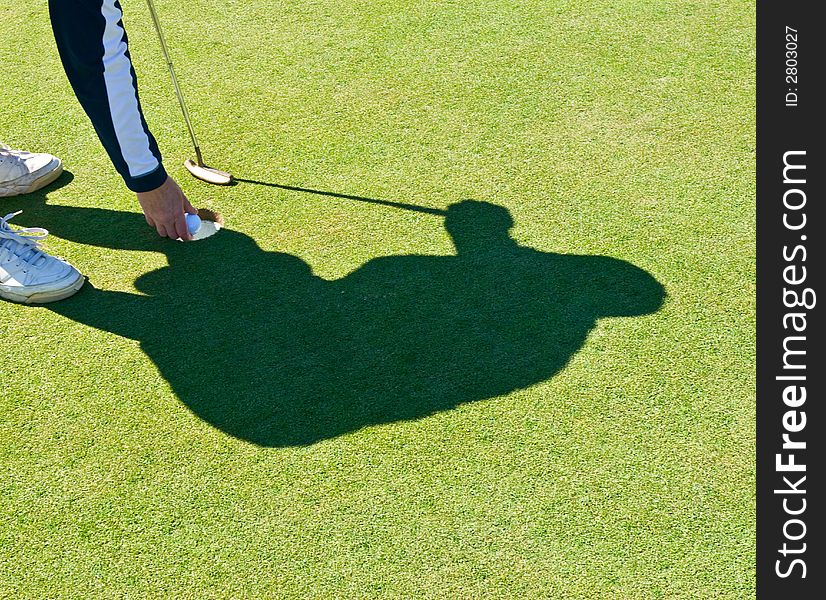 Shadow of a golfer taking the ball out of the cup after sinking a putt. Shadow of a golfer taking the ball out of the cup after sinking a putt