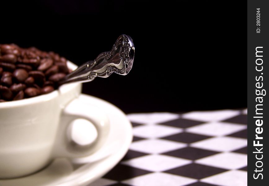 Color photo of a cup full of dark brown coffee beans with a silver spoon sticking out from it. The focus is on the tip of the spoon. Color photo of a cup full of dark brown coffee beans with a silver spoon sticking out from it. The focus is on the tip of the spoon.