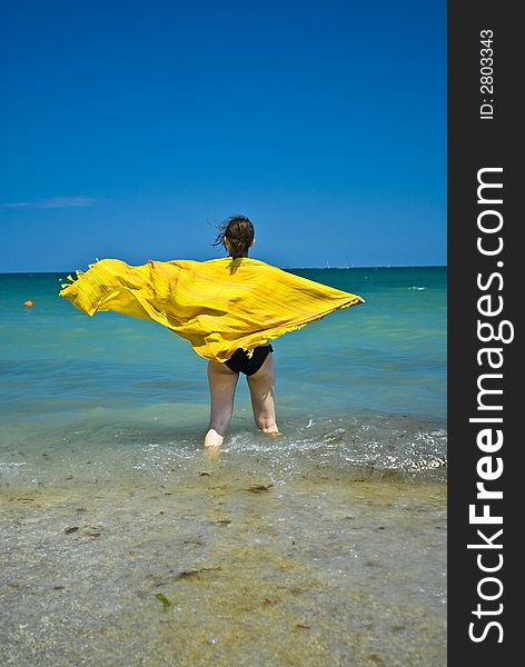 Young woman wrapped in colourful scarf blown by the summer breeze, wading in the shoreline shallow water.