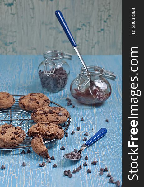 Side view of cookies with chocolate drops on round iron stand, jars with jam and chocolate on cracked blue background. Side view of cookies with chocolate drops on round iron stand, jars with jam and chocolate on cracked blue background