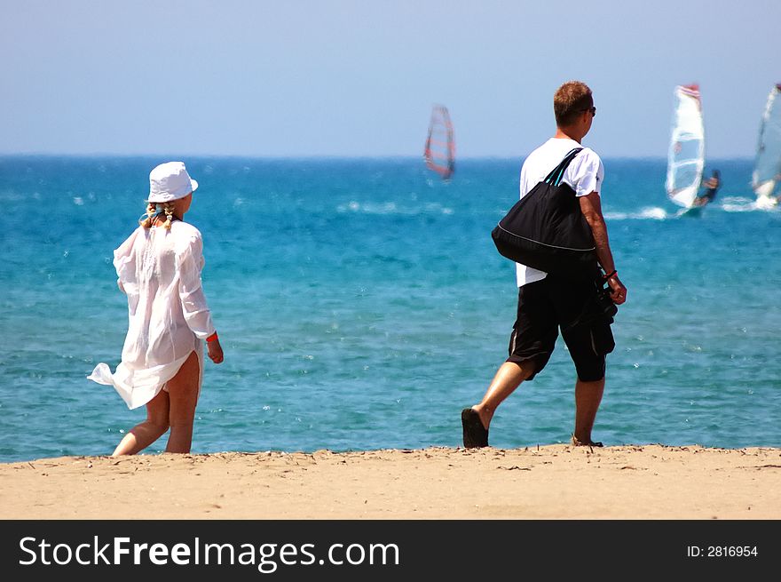 Young couple walking along a sandy beach, sunny day, summer season, in background wind surfers surfing in a blue sea. Young couple walking along a sandy beach, sunny day, summer season, in background wind surfers surfing in a blue sea.