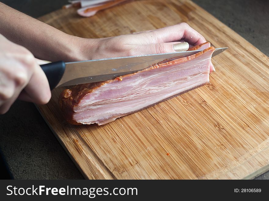 Hands slicing a big piece of bacon with a knife on a cutting board. Hands slicing a big piece of bacon with a knife on a cutting board