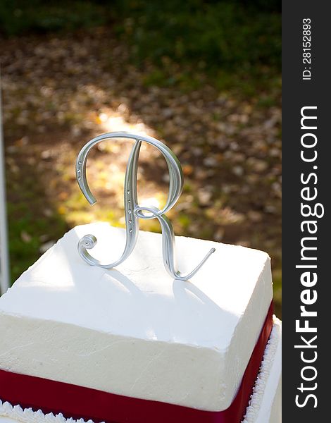 A white and red square wedding cake with multiple tiers and the letter R on top sits outside on a yellow table for a wedding reception. A white and red square wedding cake with multiple tiers and the letter R on top sits outside on a yellow table for a wedding reception.