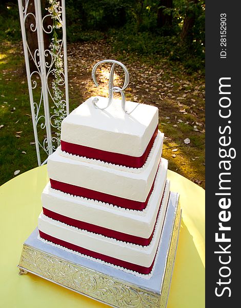 A white and red square wedding cake with multiple tiers and the letter R on top sits outside on a yellow table for a wedding reception. A white and red square wedding cake with multiple tiers and the letter R on top sits outside on a yellow table for a wedding reception.