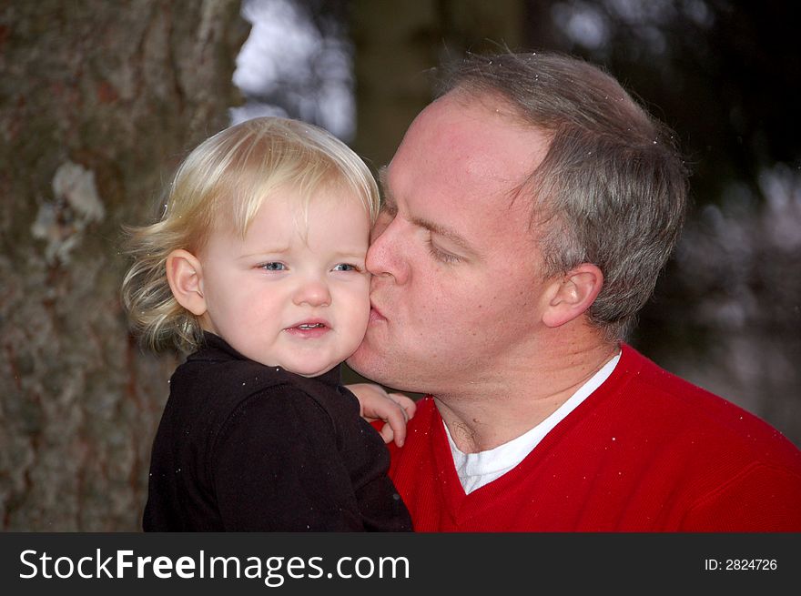 A father kissing his young daughter on the cheek.  They are outside standing in front of a tree. A father kissing his young daughter on the cheek.  They are outside standing in front of a tree.