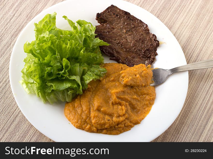 Chestnut-carrot-pumpkin mashed mix with green salad and a beef steak on the white plate. Photo is takes from the up. Chestnut-carrot-pumpkin mashed mix with green salad and a beef steak on the white plate. Photo is takes from the up.