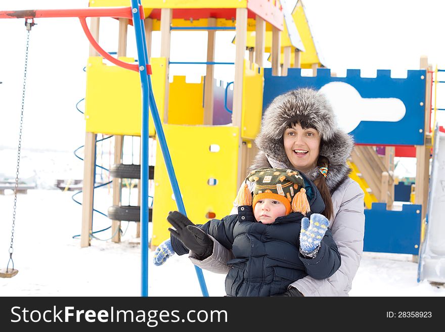 Happy mother and her young son standing in winter outfits waving at the camera in front of colourful childrens playground equipment in the snow. Happy mother and her young son standing in winter outfits waving at the camera in front of colourful childrens playground equipment in the snow