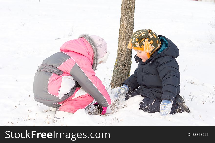 Two young children wrapped up warmly against the winter chill digging in the snow together at the foot of a small tree. Two young children wrapped up warmly against the winter chill digging in the snow together at the foot of a small tree