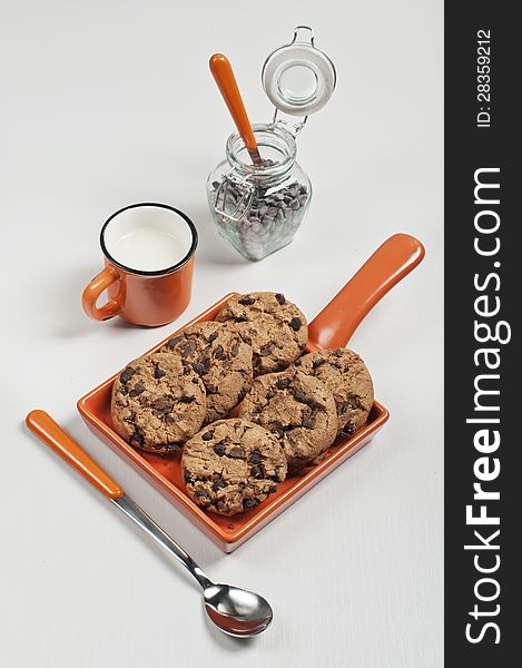 Top view of biscuits in orange ceramic pan, iar with chocolate drops, spoons and cup of milk on white wooden background. Top view of biscuits in orange ceramic pan, iar with chocolate drops, spoons and cup of milk on white wooden background