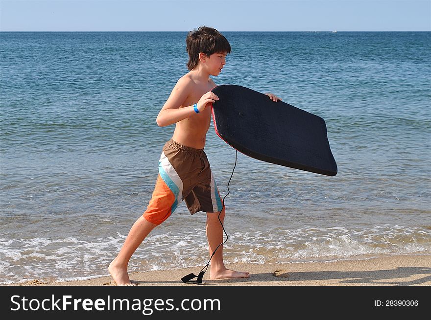 A young boy is walking in the shore at a beach with a board, ready to have great time in the water. A young boy is walking in the shore at a beach with a board, ready to have great time in the water.