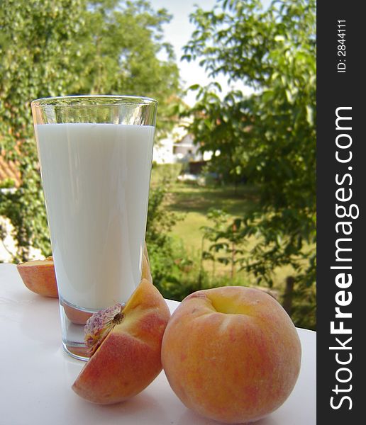 Glass of milk and a couple of peaches on a white table. Blurred scenery of the countryside in the background. Glass of milk and a couple of peaches on a white table. Blurred scenery of the countryside in the background.