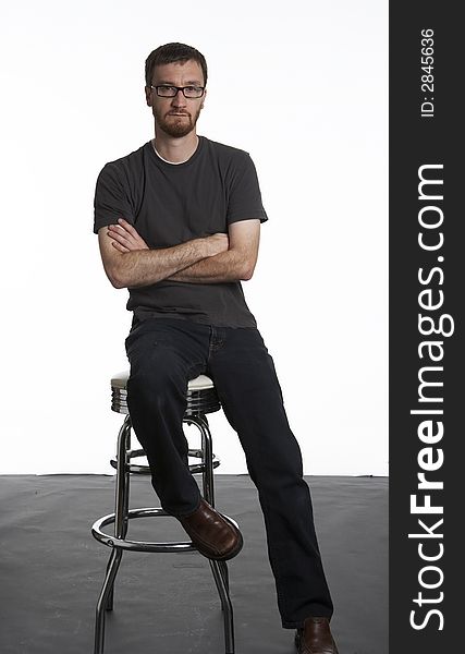 Portrait of a bearded man inside a studio. His arms are folded and he is sitting on a chrome stool. Portrait of a bearded man inside a studio. His arms are folded and he is sitting on a chrome stool