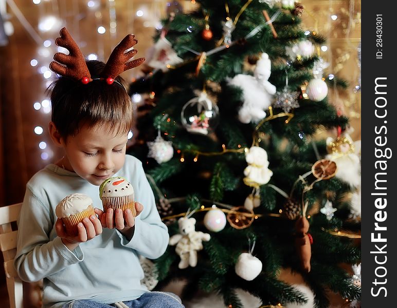 The boy with small horns of a deer under a New Year tree with beautiful fruitcakes in hand. The boy with small horns of a deer under a New Year tree with beautiful fruitcakes in hand