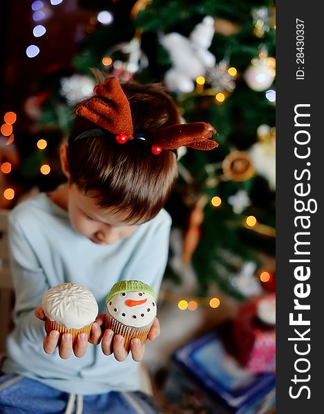 The boy with small horns of a deer under a New Year tree with beautiful fruitcakes in hand. The boy with small horns of a deer under a New Year tree with beautiful fruitcakes in hand