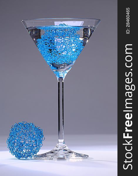 Cocktail glass formed like a cone on a stem filled with transparent fluid and a light blue mesh, in front of neutral grey background, a ball of blue mesh lying beneath it. Cocktail glass formed like a cone on a stem filled with transparent fluid and a light blue mesh, in front of neutral grey background, a ball of blue mesh lying beneath it