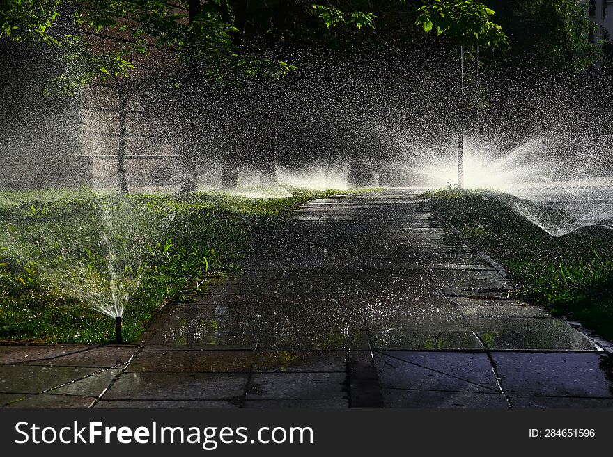 A Path Made Of Granite Slabs Over Which Water Droplets From Sprinklers Fly, Illuminated By The Morning Sun.