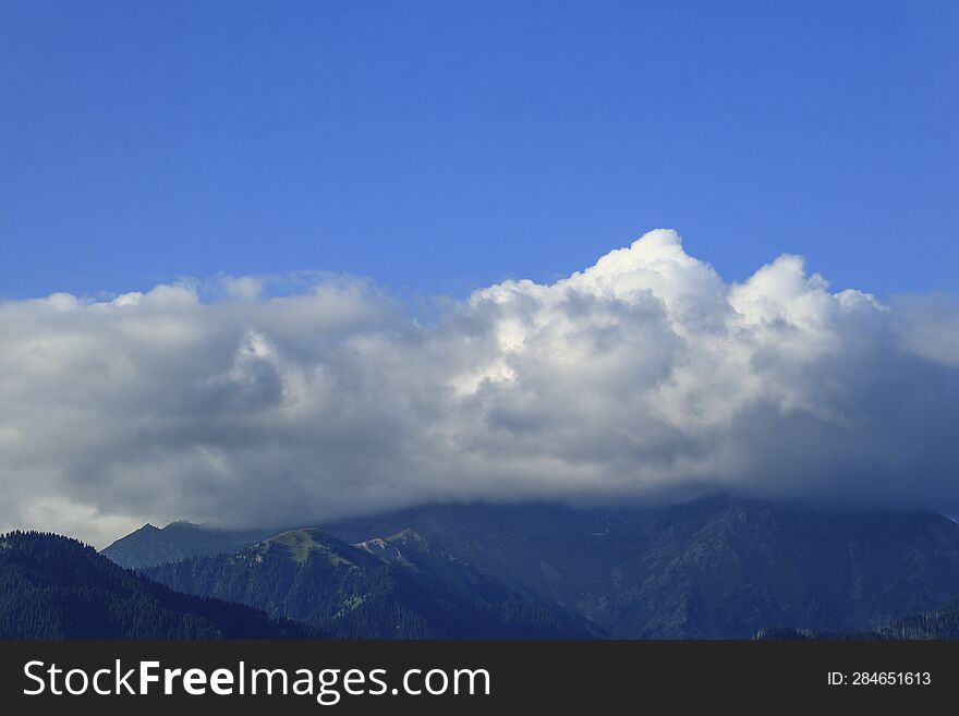 Large great white cumulus clouds in the blue sky over the mountains of the Trans-Ili Alatau, covering their peaks, are brightly li