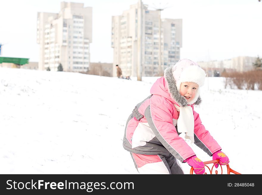 Pretty little girl in a cute furry pink outfit playing with a sled in a snowy field with two apartment blocks visible in the distance on the horizon. Pretty little girl in a cute furry pink outfit playing with a sled in a snowy field with two apartment blocks visible in the distance on the horizon.