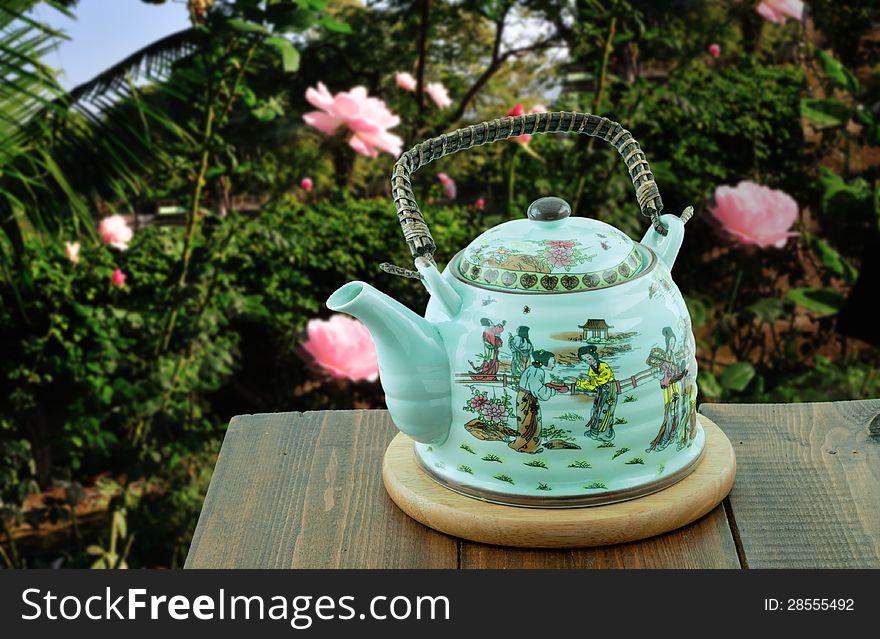 Chinese teapot standing in the garden of roses is on a wooden table on a round pedestal. Chinese teapot standing in the garden of roses is on a wooden table on a round pedestal