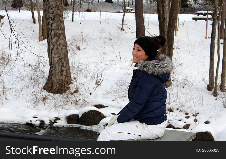 Pretty young woman with huge smile dressed warmly and sitting on stone by river's edge and fallen snow. Pretty young woman with huge smile dressed warmly and sitting on stone by river's edge and fallen snow.