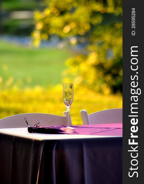 A wine glass on a table with white chairs and a purple table cloth with trees and a lake in the background