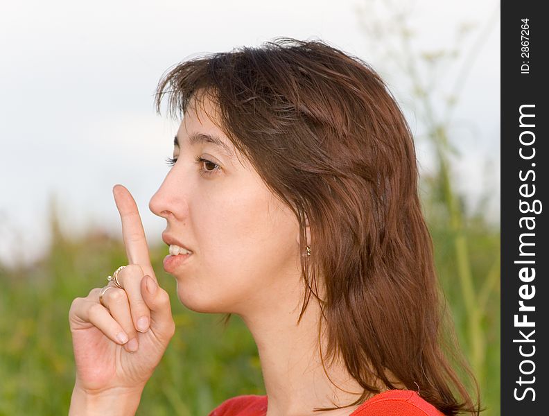 Photo of the young woman in a structure, calling for silence gesture (a finger enclosed to lips) on a background of the blue sky with clouds and a green grass. Focus of a photo on eyes of the woman